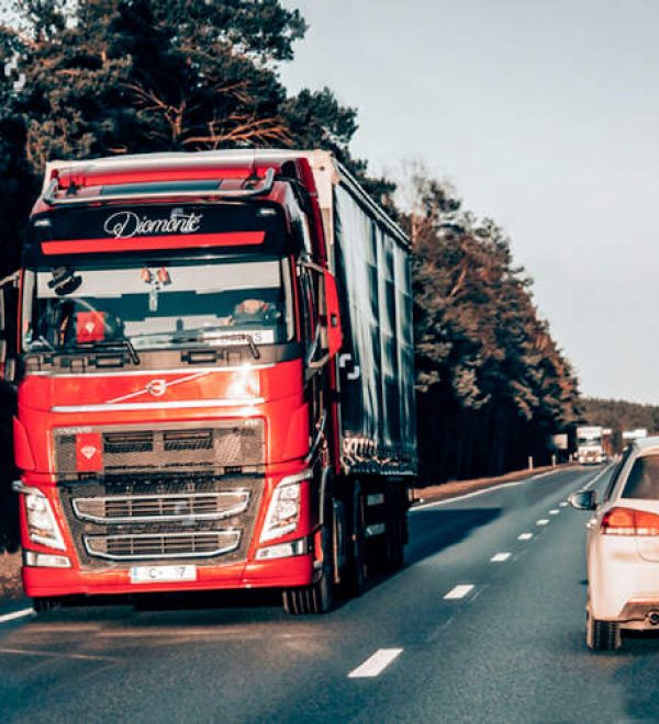 Red semi-truck and a white car sharing a two-lane highway lined with trees.